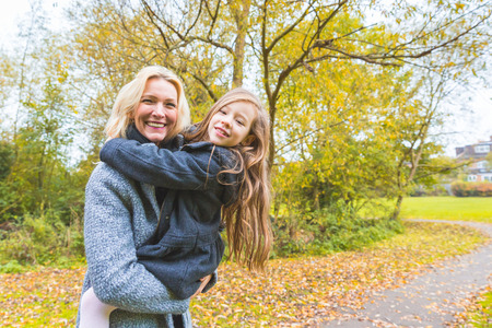 Mother and daughter portrait at park in autumn. The mother is caucasian and she is carrying her mixed race daughter on her arms. They are both looking at camera. Family lifestyle concept.の写真素材