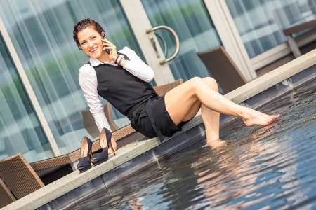 Young businesswoman having fun next to the swimming pool. She is in her mid thirties, wearing formal clothes, talking on the phone and looking at camera. Business concept.の写真素材