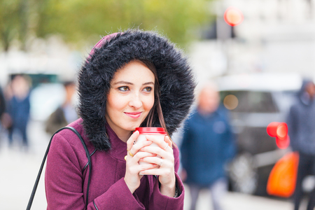 Beautiful young woman next to a busy road holding a cuf of tea. Photo taken in London, with people and traffic on background. She is wearing warm clothes. Lifestyle and loneliness concepts.の写真素材