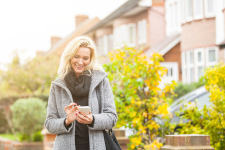 Adult woman looking at smart phone in front of some houses in London. She is a blonde woman on her early forties, she looks candid and spontaneous. Lifestyle and technology concepts.の写真素材