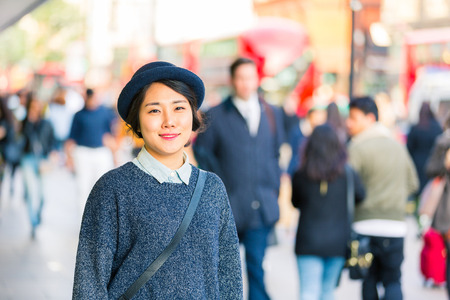 Portrait of an asian young woman with blurred people on background in a busy street of London. She is wearing a hat and looking at camera smiling. Lifestyle and travel concepts.の写真素材