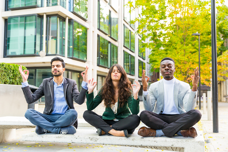 Three young business people doing yoga excercises during a break. They are two men and a woman, mixed races, sitting on a bench with modern buildings on background. Work, stress and business conceptsの写真素材