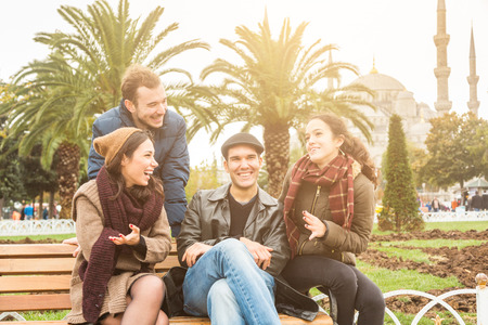 Group of Turkish friends in Istanbul. They are two men and two women, sitting on a bench at park with a mosque on background. They are wearing warm clothes. Travel and lifestyle concepts.の写真素材