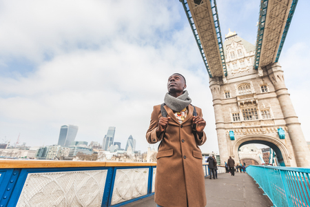 Young black man standing on Tower Bridge with London skyline  on background. He wears a brown coat and he is looking away from camera.の写真素材