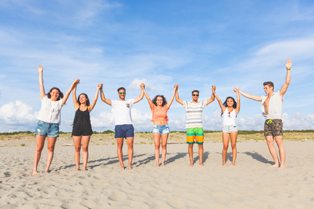 Multiracial group of friends with raised hands on the beach. There are four girls and three boys, wearing trunks and colourful t-shirts. Achievement and success concepts.の写真素材