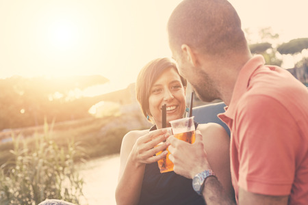 Couple having a cocktail and toasting outdoor on summer. The bar is next to a river, backlight scene at sunset. They are looking each other, the man is showing his shoulders to the camera.の写真素材