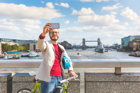 Hipster man taking a selfie in London with Tower Bridge on background. He is commuting, he has a fixed gear bike and holds the helmet with the arm. Lifestyle, travel and sustainability concepts.の写真素材