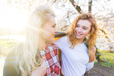 Two lesbian girls at park in London, looking each other while walking. They are holding a large scarf on their shoulders to keep warm on a sunny spring day. Homosexuality and lifestyle concepts.の写真素材