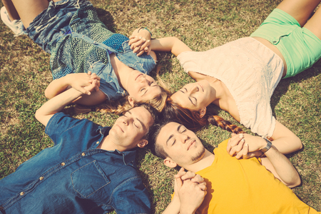 Friends lying and relaxing on the grass . They are two girls and two boys resting at park, holding hands and keeping eyes closed. Beautiful image of friendship and togetherness.の写真素材