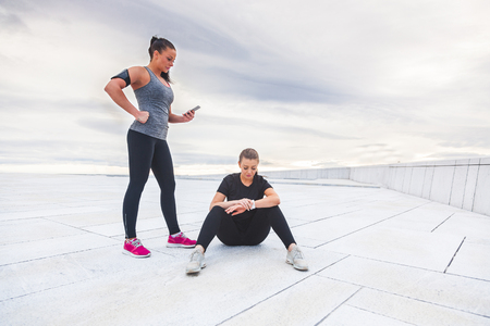Two women checking workout statistics on their electonic devices. They are wearing gray and black sportswear and resting after fitness exercises. Technology and health lifestyle conceptsの写真素材