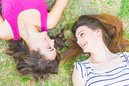 Two girls lying on the grass and laughing looking each other. One has curly hair and the other has streight hair, both brunette. Relaxation and friendship concepts.の写真素材