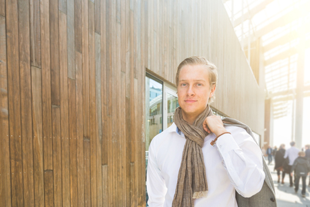 Fashioned young businessman portrait. He has nordic facial features, wearing a white shirt and holding light gray blazer. Backlight with flares and some blurred people on background.の写真素材