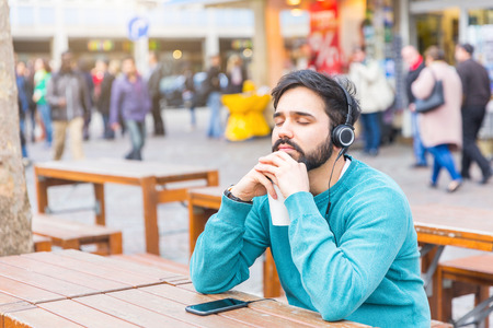Young man relaxing and listening to music with headphones in the city. He is keeping his eyes closed while holding a cup of coffee. There are blurred people on background.の写真素材