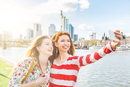 Two beautiful young women taking a selfie with Frankfurt skyline on background on a spring day. They are smiling and looking at the smart phone. Friendship and travel concepts.の写真素材