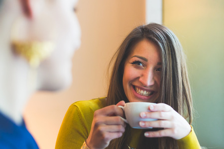 Two women in a cafe smiling and looking each other. One is blurred on the left and the other woman is looking at her, smiling and holding a cup of coffee. She looks candid with a beautiful smile.の写真素材