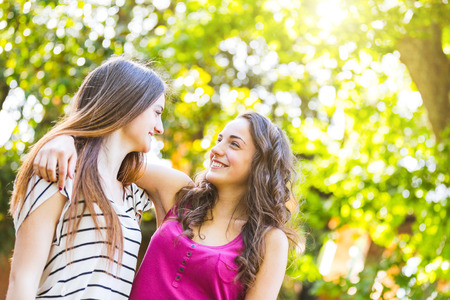 Two girls embraced together at park. They are happy and smiling while looking each other. Best friends embracing with trees on background. Lifestyle and friendship concepts.の写真素材
