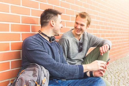 Two men sitting next to a brick wall, holding a smart phone and looking each other with a smiling face. Urban city life situation with real people. Friendship and lifestyle concepts.の写真素材