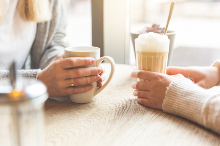 Two beautiful young women in a cafe, drinking coffee and latte macchiato. Close up shot on the hands holding the mugs.の写真素材