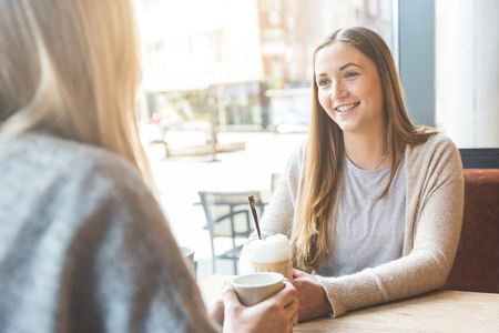 Two beautiful young women in a cafe, drinking coffee and latte macchiato and talking. They are sitting in front of each otherの写真素材