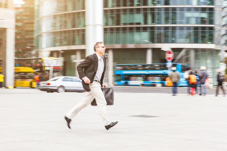 Businessman running in the city. Blurred background with panning technique. Caucasian man on his late fifties, wearing smart casual clothes and holding a suitcase. Business and city life concepts.の写真素材