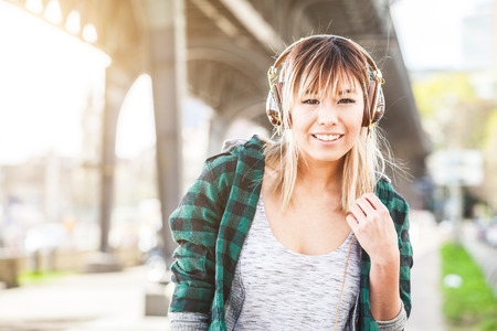 Portrait of a beautiful young woman in Hamburg listening music. She is Japanese, on her early twenties, brunette, wearing big headphones and listening music, looking at camera and smiling.の写真素材