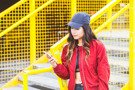 Asian woman typing on her smart phone in London. She is New Zealander, with asian face features, on her mid twenties and she is looking at the phone and typing. Urban style clothes.の写真素材