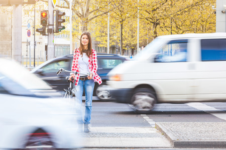 Young woman with bicycle waiting to cross the street at red signal in Berlin. There are blurred cars passing in front of her and behind. Urban lifestyle and travel concepts.の写真素材