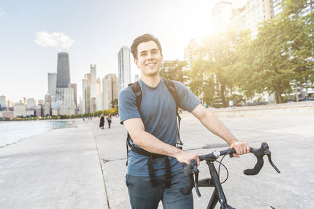Man walking carrying his bike in Chicago. Young caucasian man looking at the camera, smiling. Downtown skyscrapers on background. Urban outfit, travel and youth culture concepts.の写真素材