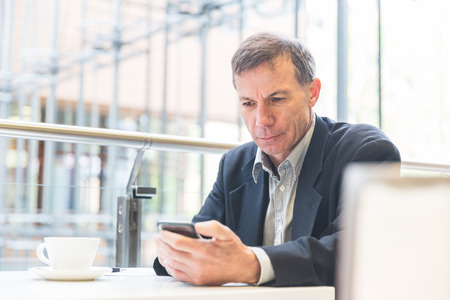 Businessman looking at the smart phone in a cafe. Caucasian man on his late fifties, typing on his smart phone. There is a coffee on the table. Business and lifestyle concepts.の写真素材