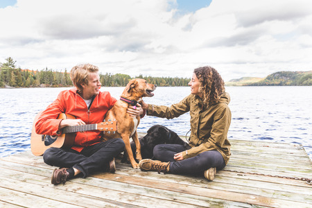 Happy young couple playing with their dog on a dock. The man is holding a guitar, they are wearing a rain jacket, love and nature conceptsの写真素材