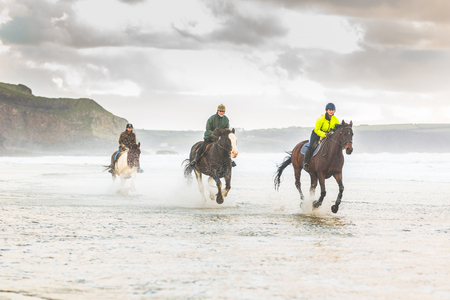 Horses galloping on the beach. Three people riding horses at seaside on a stormy day. Epic photo of horses on the sea with water splashing. Sport and travel conceptsの写真素材