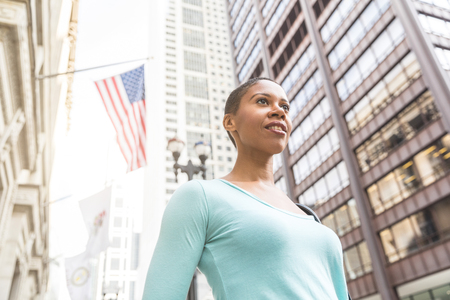 Woman portrait with USA flag on background. Proud black woman in Chicago downtown, looking away from camera. The woman has short hair and wears casual clothesの写真素材