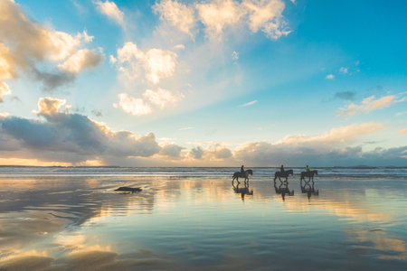 Horses walking on the beach at sunset. Three people riding horses at seaside on a cloudy day. Epic photo, wide angle shot, backlight with silhouette. Sport and travel conceptsの写真素材