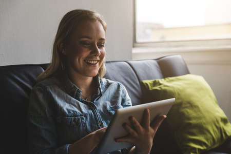Young woman typing on a digital tablet at home. Beautiful girl on the sofa, sitting and holding the electronic device. Smiling happy face, lifestyle and technology concepts.の写真素材