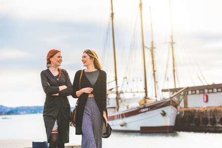 Two beautiful girls walking at Oslo harbour enjoying life talking and looking each other with ships on background Lifestyle and friendship conceptsの写真素材