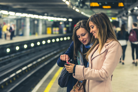 Couple of female friends looking at the smartphone while waiting for the train on the tube. Two young women checking on the phone before boarding. Tone addedの写真素材