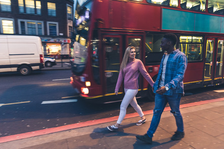 Mixed race couple walking in London at night. Multiracial couple hanging out in the city, panning view with red bus and traffic on background. Lifestyle and travel conceptsの写真素材