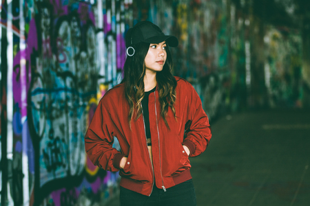 Asian woman portrait with blurred graffiti on background Beautiful young woman listening music with headphones. Urban style clothes and backgroundの写真素材