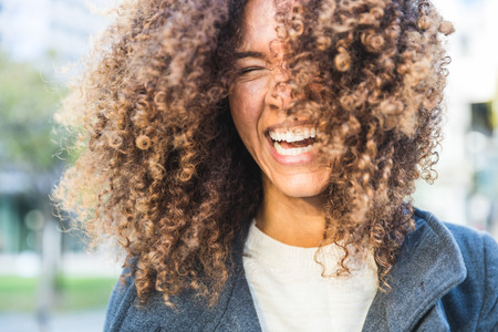 Curly woman laughing and shaking head. Smiling mixed race woman with curls having fun. Smart casual dress. Lifestyle and hairstyle conceptsの写真素材