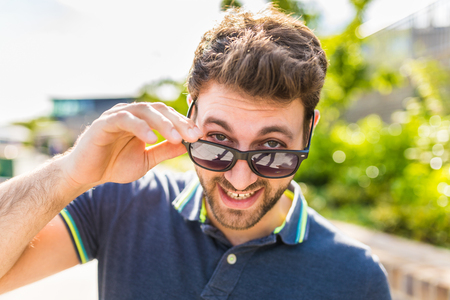 Happy smiling man portrait at park. Young man holding sunglasses and looking at camera. Cheerful caucasian face with brown beard and hair.の写真素材