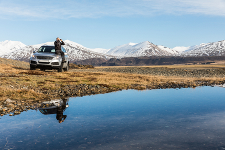 Man explorer in Iceland sitting on the car. Adult man on the bonnet looking through binoculars. Majestic mountains on background, reflections on water on foreground. Travel and wanderlust conceptの写真素材