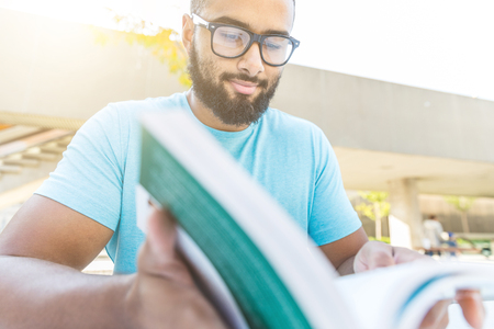 Black man reading a book in the city. Young man with long beard, wearing black eyeglasses and a light blue t-shirt sitting at a table with a book. Lifestyle and relax conceptsの写真素材