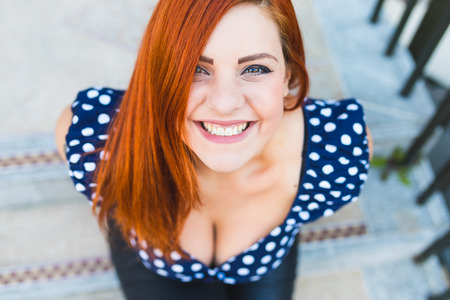 Redhead woman portrait. Smiling girl looking up at camera, wearing a dotted pin up style top and sitting on concrete steps. Happiness and carefree conceptsの写真素材
