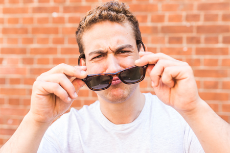 Funny man with sunglasses portrait wall on background. Young man holding sunglasses and looking at camera. Cheerful caucasian man wearing white t-shirtの写真素材