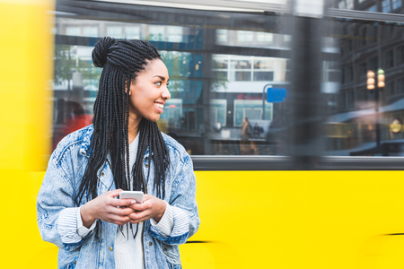 Black girl typing on smart phone in Berlin. Beautiful young woman in the city with a blurred tram in Alexanderplatz on background. Lifestyle, technology and travel conceptsの写真素材
