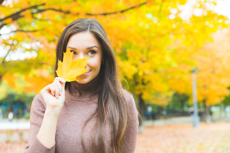 Portrait of a beautiful young woman hiding behind a yellow leaf. Smiling woman wearing turtleneck sweater with trees on background. Autumn season theme.