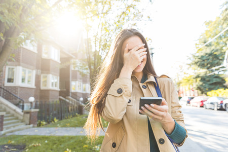 Woman with smartphone laughing and covering face with hand. Beautiful and happy girl wearing beige trench coat having fun in the cityの写真素材