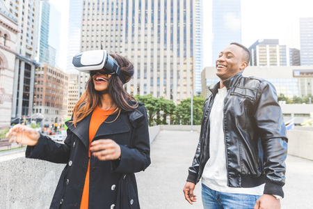 Black woman trying virtual reality headset and laughing. A man is looking at her and smiling. They...