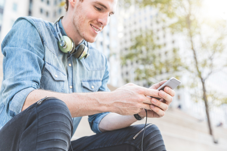 Man with phone and headphones in Chicago. Portrait of a ginger young man holding a smartphone and typing. Buildings and trees on backgroundの写真素材