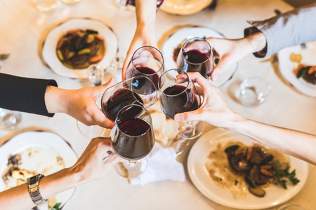 People toasting with red wine at restaurant. Close up view of five hands holding glasses with wine, and some food on the table. Lifestyle and drinks concept with an Italian style.の写真素材